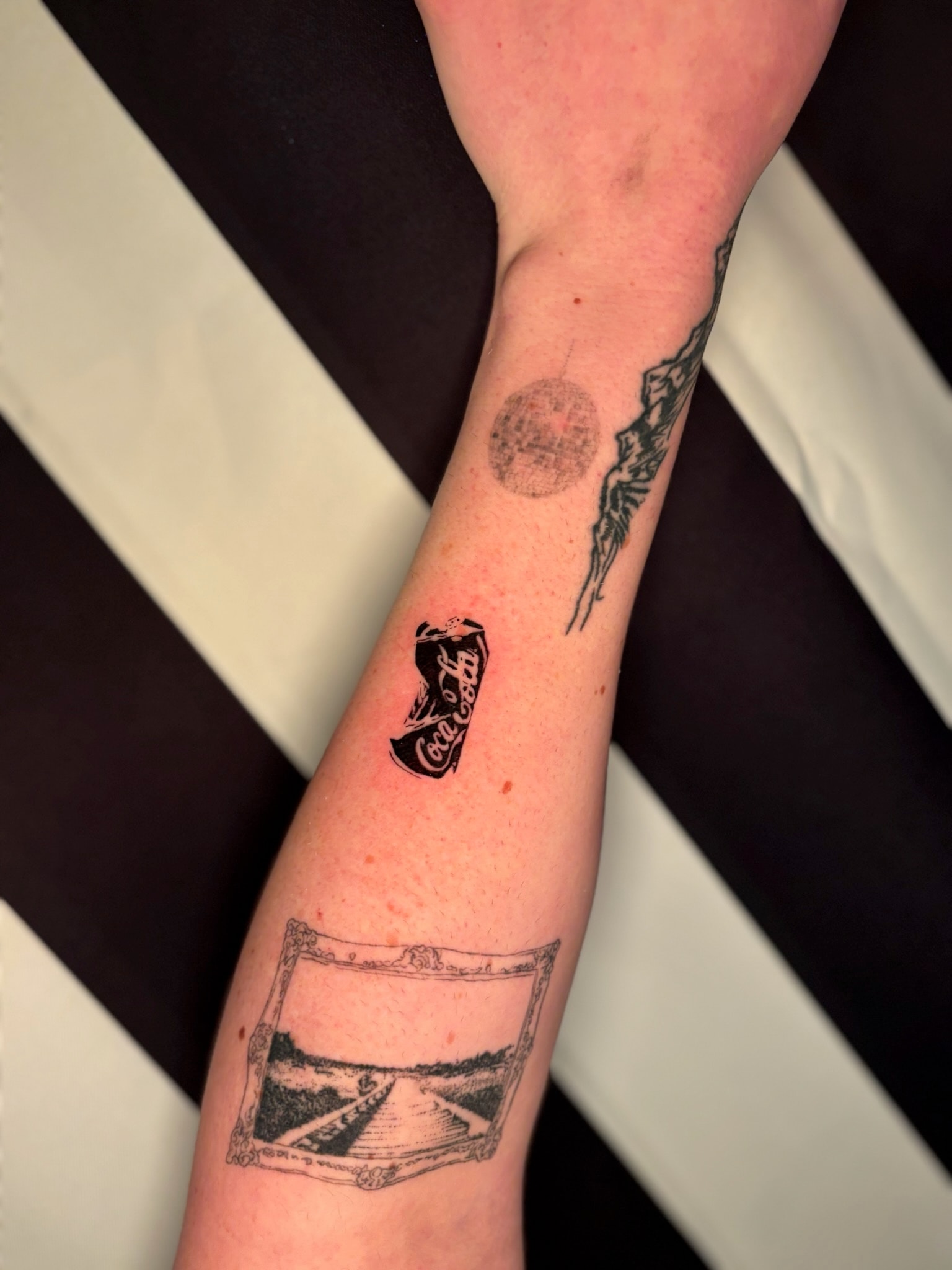 Fine line arm tattoos including crushed Coca-Cola can, disco ball, and framed railroad landscape