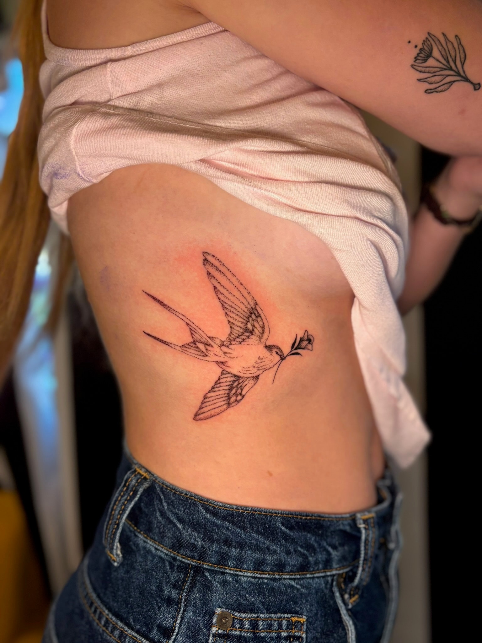 Fine line swallow bird carrying a flower on ribs, black and grey fine line